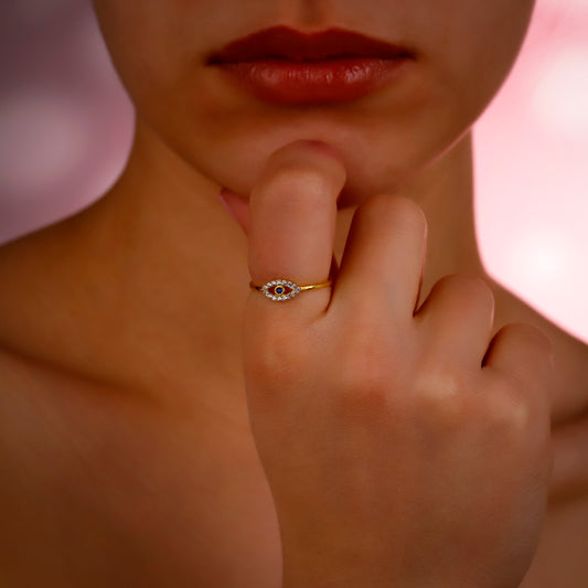 Close-up of a hand with a gold ring featuring an eye design, against a soft pink background.