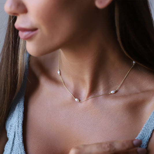 A close-up image of a woman wearing a gold choker necklace with freshwater pearls, over a light blue spaghetti strap top.