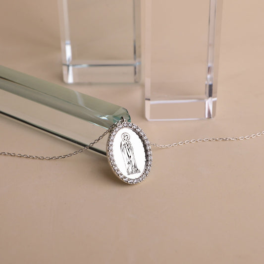 A silver necklace featuring a round pendant with an image of the Virgin Mary, encapsulated by cubic zirconia crystals, displayed on a flat surface.