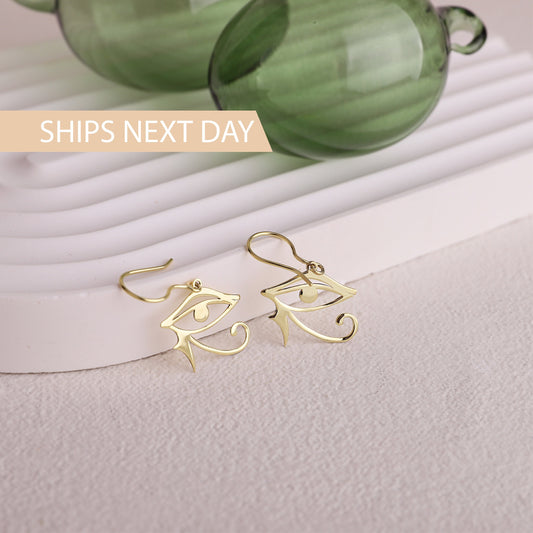 Gold eye-shaped earrings on a light surface with green glass objects in the background.