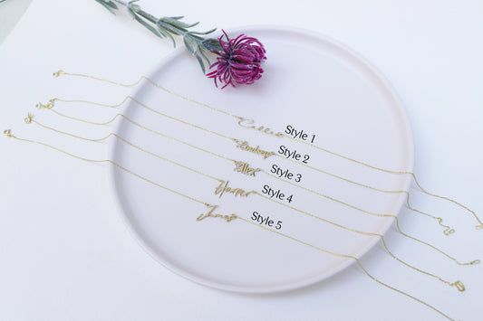 A close-up image of some gold-filled nameplate necklaces with the name written in a different cursive fonts on a chain.
