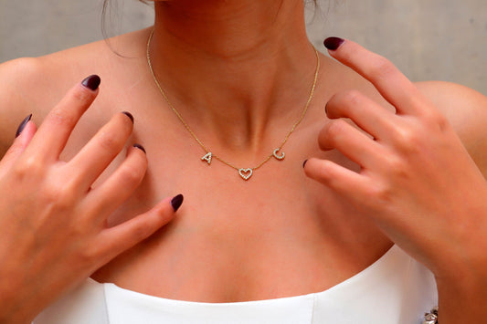 A close-up image of a woman's neck wearing a dainty two-initial pave necklace with a heart-shaped charm.