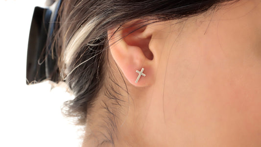 A close-up image of a person's ear wearing a 14K gold stud cross earring, with the earring's reflection visible. The person has their eyes closed and is touching the earring gently.