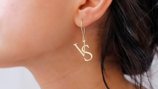 A close-up image of a woman's ear wearing a dangle earring with the initials 'V' and 'S'.