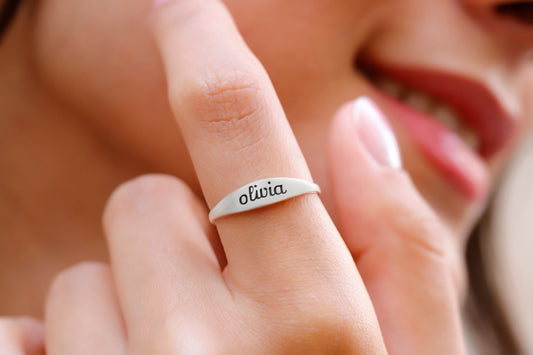 A close-up image of a woman's finger wearing a silver stackable ring with an engraved name.