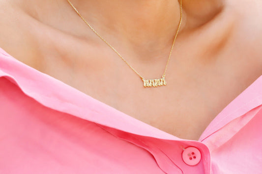 A close-up image of a woman wearing a goldcolored necklace with a 3D balloon-shaped pendant that reads 'mama'.