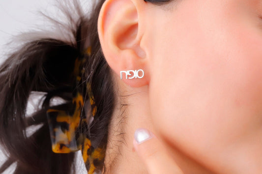 A close-up image of a woman's ear wearing a small gold-colored stud earring with a personalized Hebrew name.
