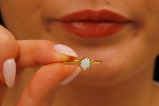 A close-up of a woman's finger wearing a gold band ring with a white opal center stone, held between her lips.