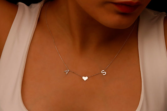 A woman wearing a white tank top showcasing a delicate minimalist necklace with initial letters and a heart charm.