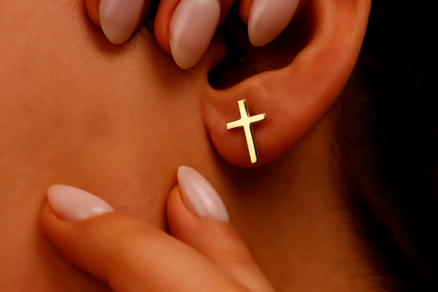 Gold cross earring on a person's ear with a hand holding the earlobe.