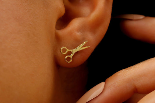 A close-up image of a person's ear wearing a small gold-colored scissor stud earring.