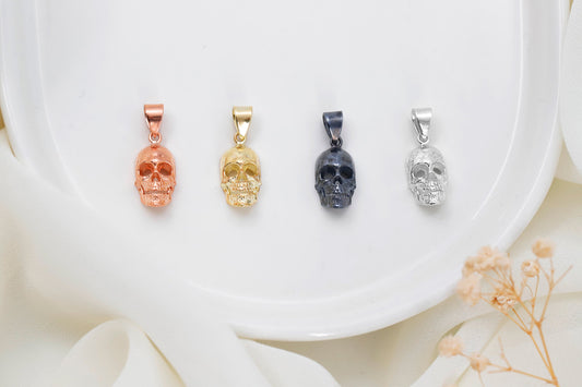 A collection of four skull-shaped charms in different finishes (oxidized silver, gold, rose gold, and plain gold) displayed on a white plate with flowers around it.