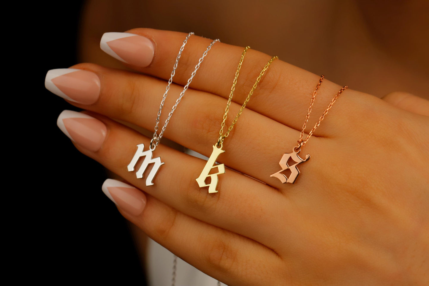 A close-up image of a woman holding 3 necklaces with a gothic style initial pendants in gold rose and silver.