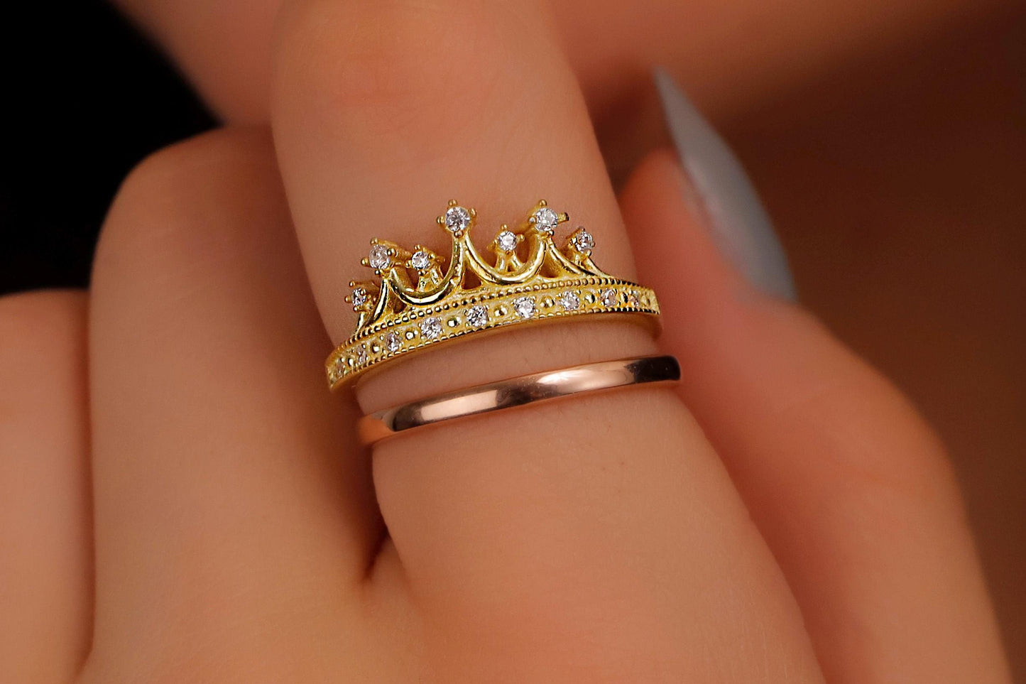 A close-up image of a woman's hand wearing two rings, one with a gold crown design and another with a rose gold band.