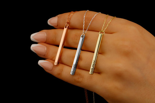 Three necklaces with long bars in silver, gold, and rose gold colors, held between fingers with a dark background.