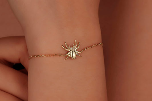 A gold-colored bracelet featuring a realistic spider design on a chain, displayed on a person's wrist.