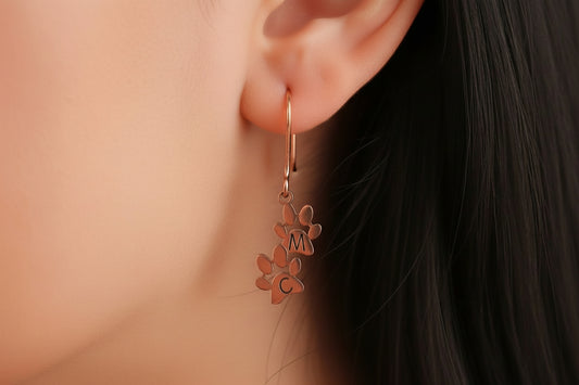 A pair of custom initial letter earrings, with one earring featuring a paw print, engraved with the letter 'M', made of sterling silver, displayed on a person's ear.