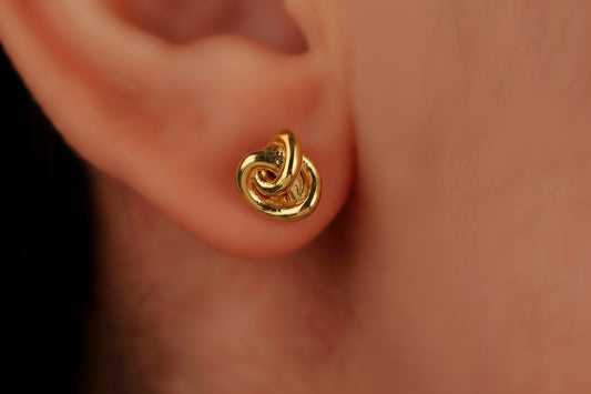 A pair of small dainty stud earrings with a love knot design, held between fingers, showcasing the jewelry's delicate and minimalist design.