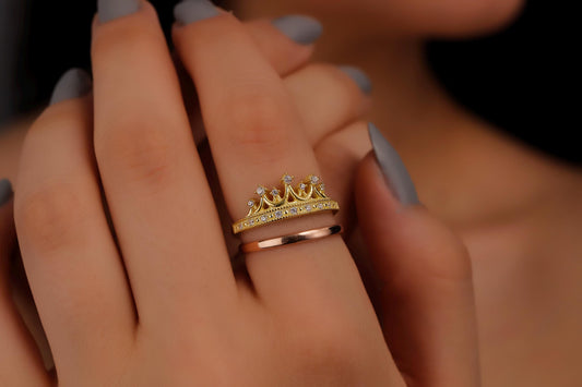 A close-up image of a woman's hand wearing two rings, one with a gold crown design and another with a rose gold band.