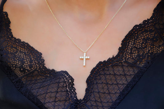 A close-up image of a person wearing a 14K gold diamond cross necklace with a delicate chain, displayed against a lacy black background.