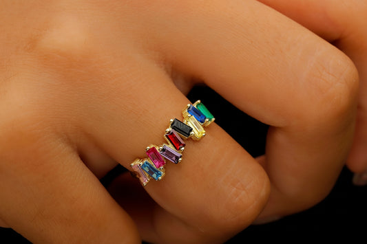 A close-up image of a person's hand wearing a multicolored baguette cut gemstones ring, with a variety of stones set in a gold-colored band.