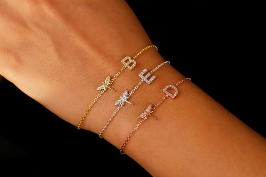 A series of three bracelets worn on a person's wrist, featuring charms with different designs, including a dragonfly and various letters.