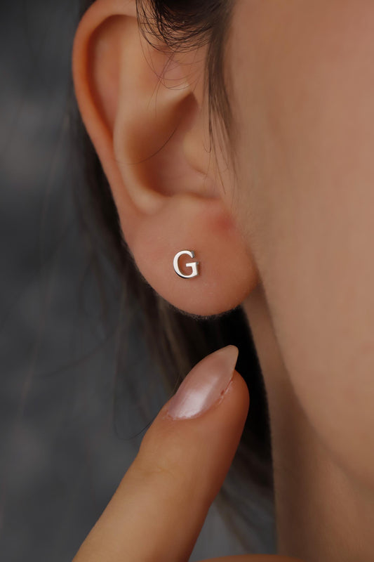 Silver 'G' earring on a person's ear with a blurred background