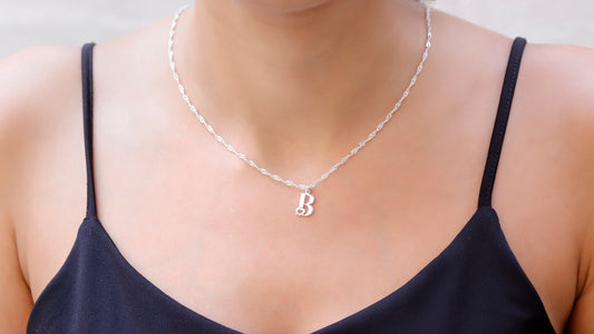 A person wearing a black tank top showcasing a white gold ball chain necklace with a single initial 'B' and a tiny heart pendant.