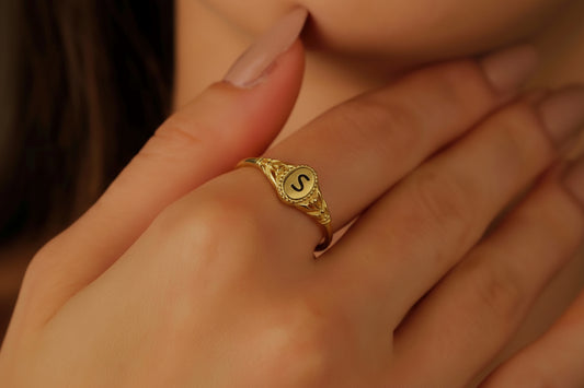 A close-up image of a person's hand wearing a gold-colored ring with an engraved initial.