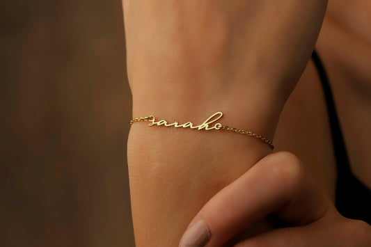 A gold bracelet with the name 'Sarah' written in cursive script on a person's wrist.