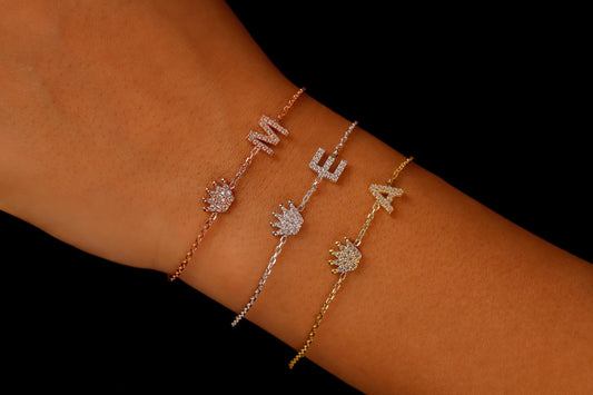A series of three dainty bracelets worn on a wrist, featuring initials and crown charms with cubic zirconia stones.