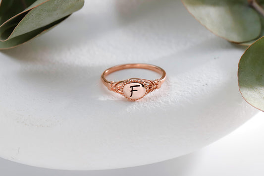 A rose gold initial ring with the letter 'F' inscribed, placed on a white surface with green leaves in the background.