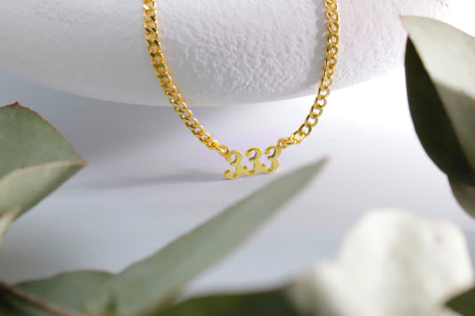 A gold-colored necklace with the numbers '333' displayed, placed on a white surface with green leaves in the background.