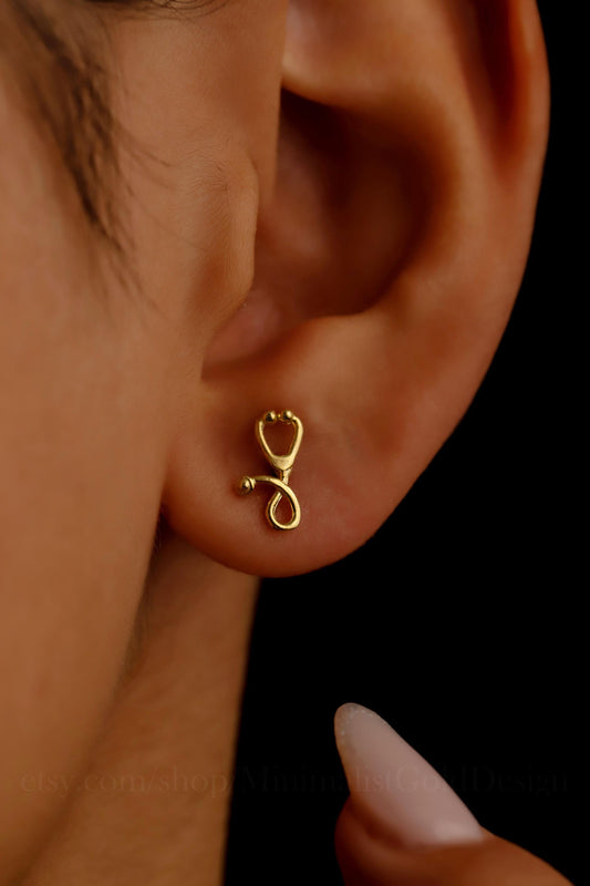 A close-up image of a person wearing a gold-colored stethoscope-shaped earring in their ear.