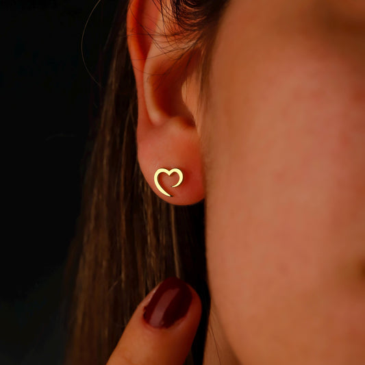 Close-up of an ear wearing a gold heart-shaped earring with a blurred background.