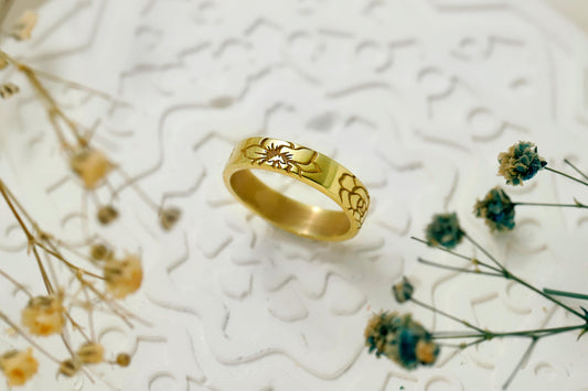Gold ring with intricate design on a textured white surface with dried flowers.
