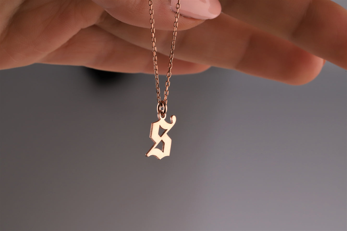 A close-up image of a woman holding a rose gold necklace with a gothic style initial pendant.