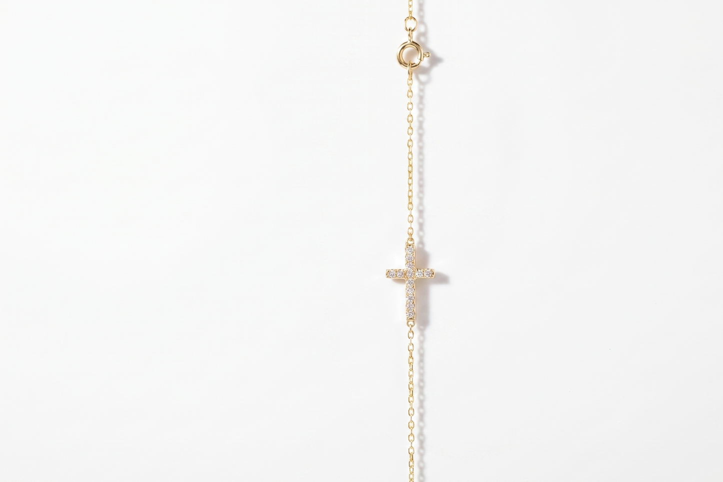 A delicate gold chain necklace featuring a small cross pendant and a tiny round bead at the top, displayed against a solid white background.
