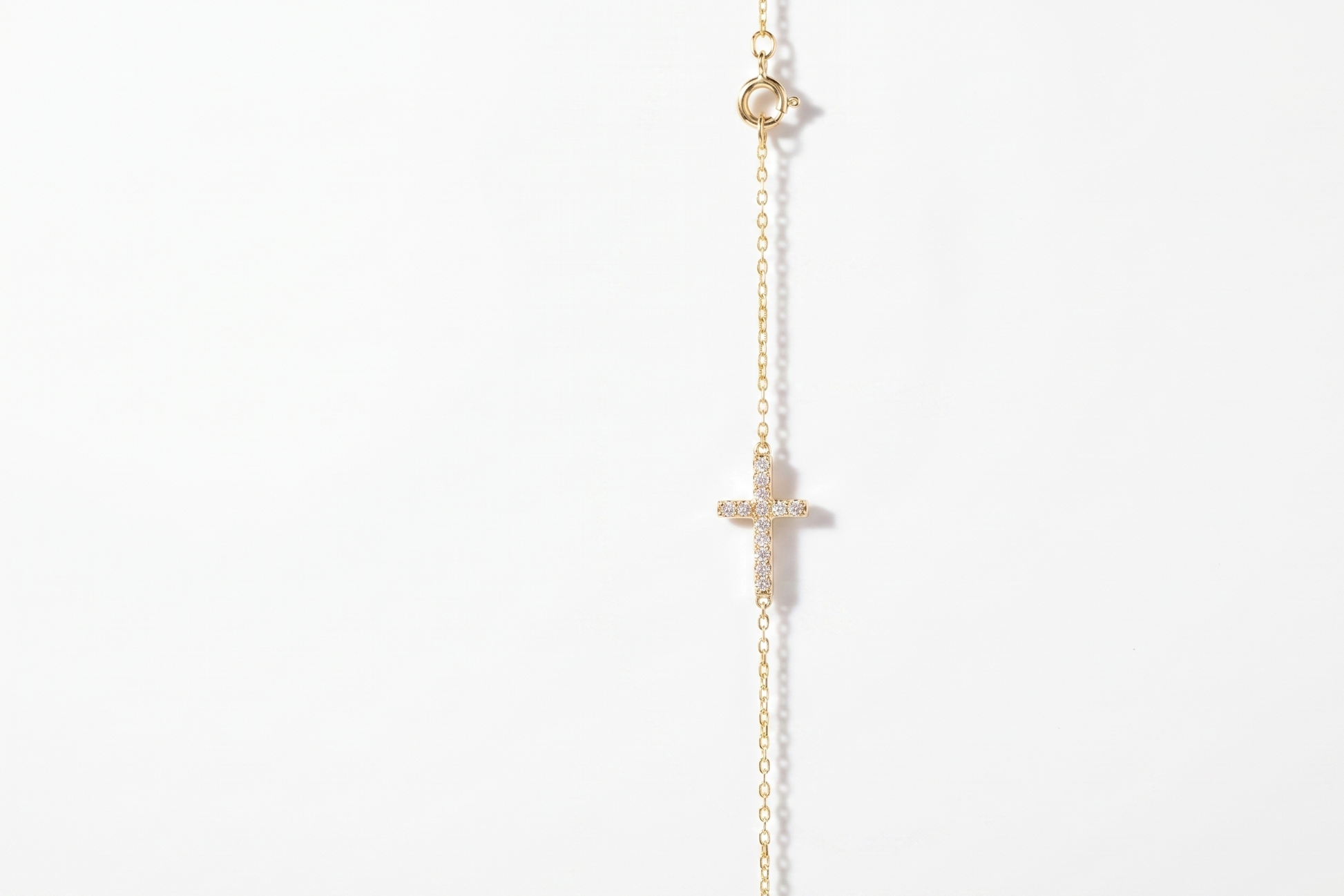 A delicate gold chain necklace featuring a small cross pendant and a tiny round bead at the top, displayed against a solid white background.