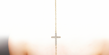 A delicate gold cross pendant necklace with small clear stones, worn on the back of a woman's neck over a black strap top, against a bright white background.