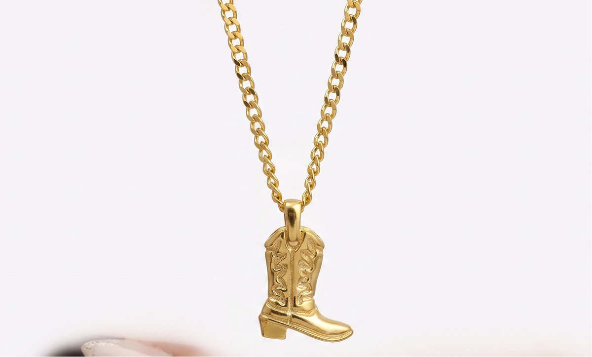 A gold chain necklace with a cowboy boot pendant, held above an open hand with manicured nails against a white background.