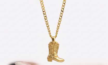A gold chain necklace with a cowboy boot pendant, held above an open hand with manicured nails against a white background.