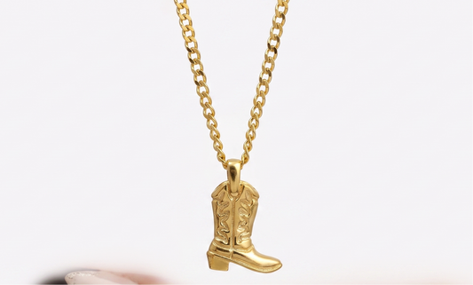 A gold chain necklace with a cowboy boot pendant, held above an open hand with manicured nails against a white background.