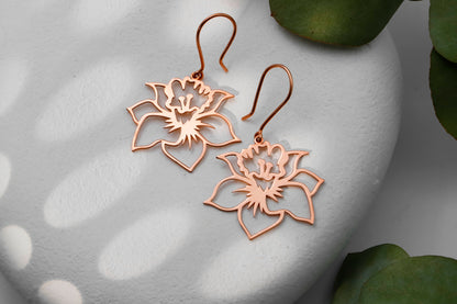 Three birth flower earrings in different colors (gold, silver, rose gold) displayed on a white surface with a green leaf in the background.