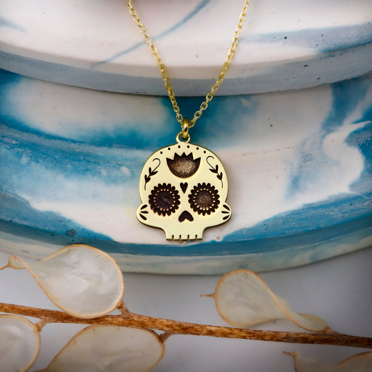Gold skull pendant necklace on a blue and white ceramic dish with dried flowers.
