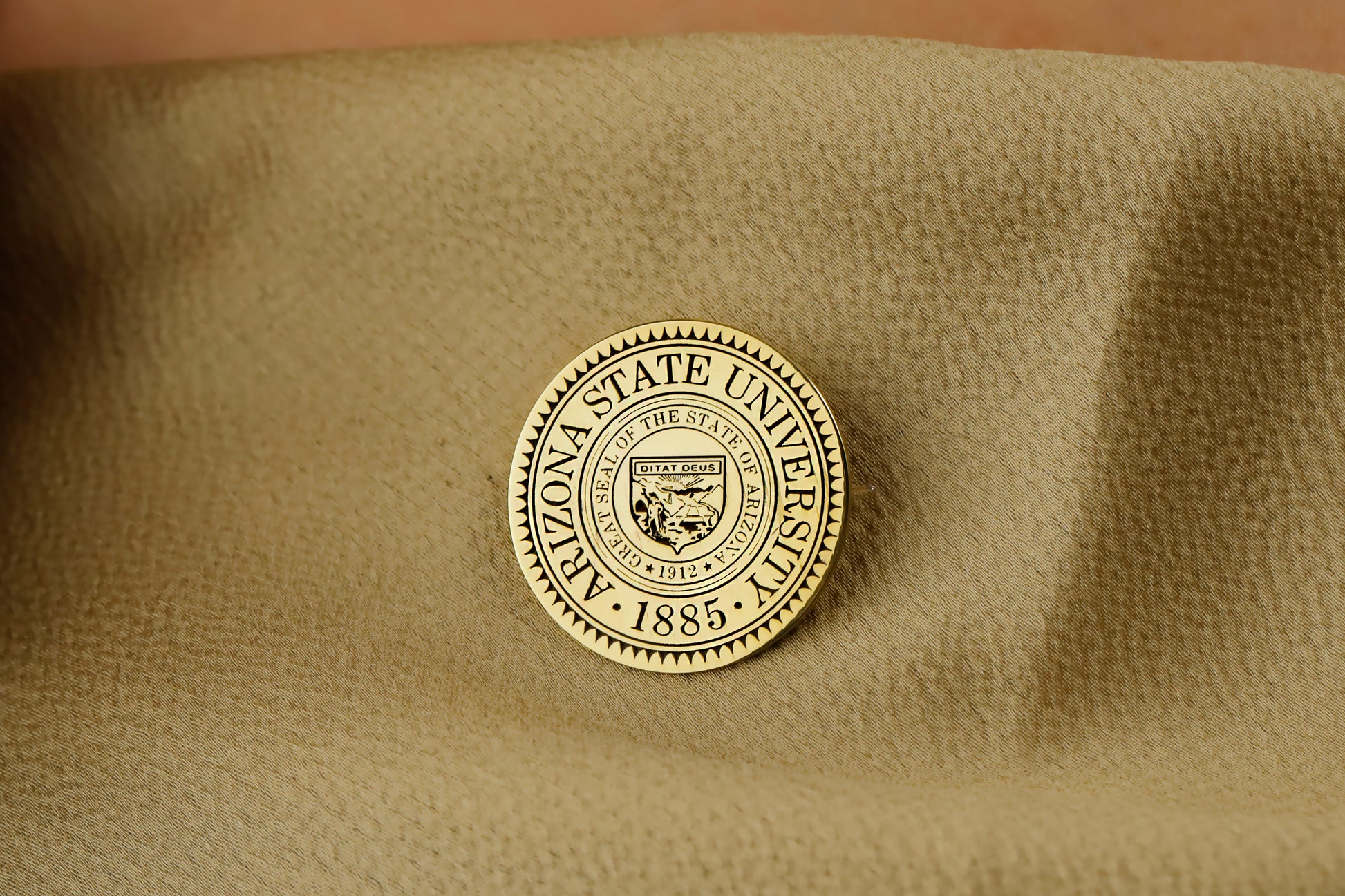 A gold-colored brooch with an engraved university seal placed on a white fabric with a floral pattern.