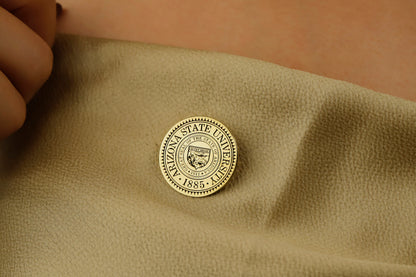 A gold-colored brooch with an engraved university seal placed on a white fabric with a floral pattern.