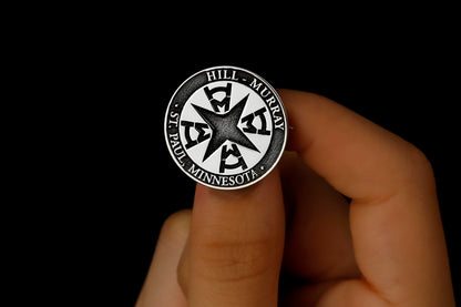 A silver brooch with an engraved university seal placed on a white fabric with a floral pattern.