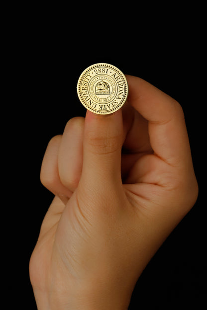 A gold-colored brooch with an engraved university seal placed on a white fabric with a floral pattern.