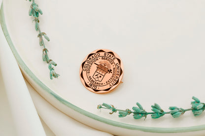 A rose gold brooch with personalized college emblems, displayed on a white surface with a green floral decoration.